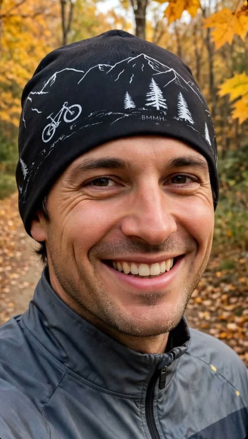 black cycling beanie with gray graphics of bicycle, mountains, pine trees, and topographic lines on a man in the forest with autumn background