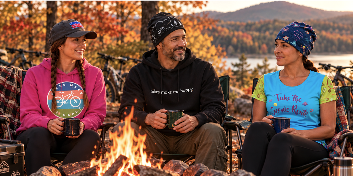 2 women and a man sitting around a campfire in a fall setting with leaves changing color. They are wearing cycling lifestyle clothing and hats. 