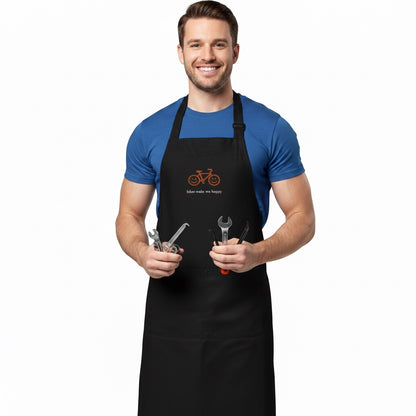 A man wearing a black Bikes Make Me Happy organic cotton apron with a smiley bike logo, holding bicycle tools.