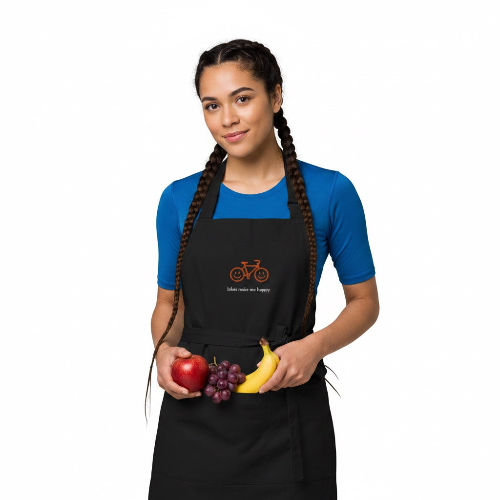 Woman wearing a black Bikes Make Me Happy organic cotton apron with a smiley bike graphic while holding fresh fruit